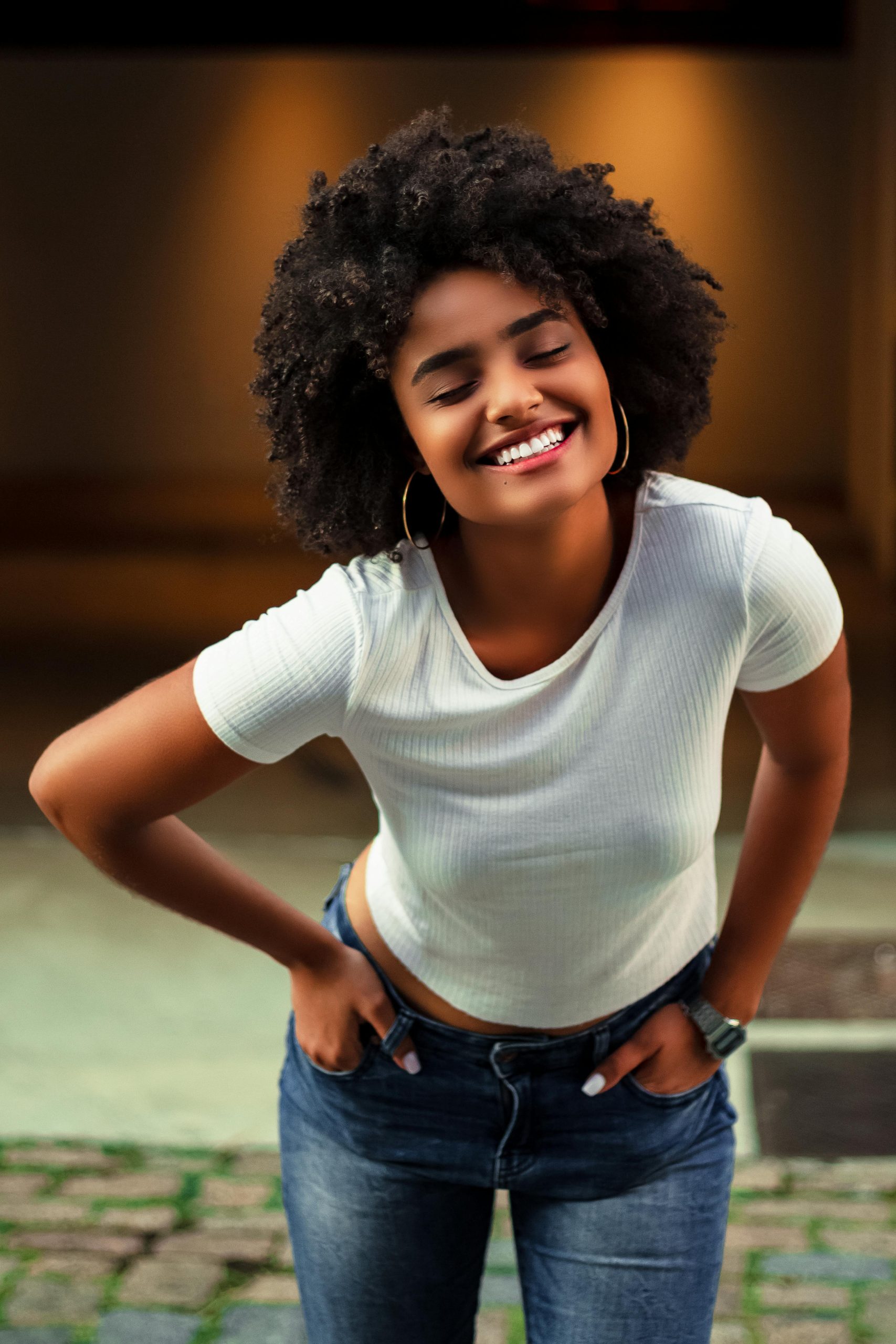 Home African American woman with afro hair smiling in casual white shirt and jeans, standing outdoors.